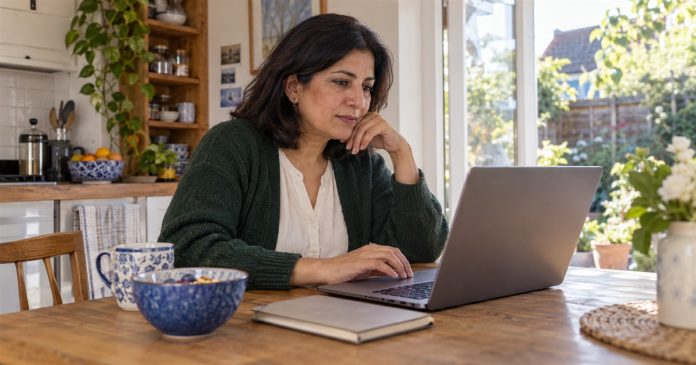 Woman in a forest-green cardigan comparing information on a laptop at a home kitchen table