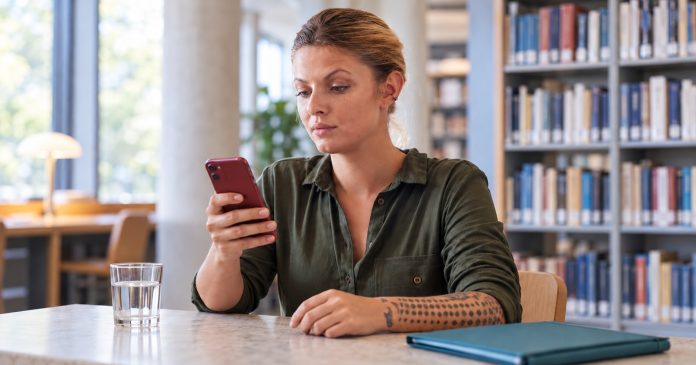 A woman checking messages on a red smartphone at a clear library table beside a glass of water and a closed teal folder.